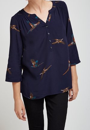 Granatowa bluzka z długim rękawem, zapinana na guziki dekolt w szpic i kolorowe nadruki ptaków, noszona z czarnymi spodniami, ukazująca górną część tułowia i ramiona.
