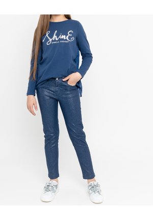 Mädchen trägt ein blaues Langarmshirt mit dem Schriftzug "Shine Sparkle Everyday", funkelnde blaue Hosen und weiße Sneakers mit Blumenverzierungen.