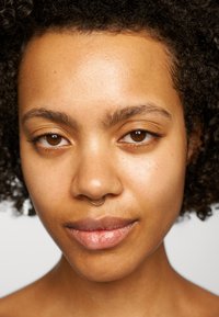 Nahaufnahme eines Gesichts mit lockigem, dunklem Haar, glatter Haut, einem goldenen Septumring und braunen Augen. Natürlichem Make-up und minimaler sichtbarer Reflexion.