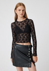 Schwarzes Spitzen-Longsleeve-Croptop mit Blumenmuster und Wellenrand, kombiniert mit einem hochgeschnittenen schwarzen Leder-Minirock mit Druckknöpfen.