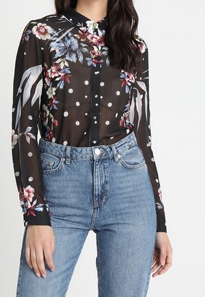 Blusa negra transparente con patrones florales y lunares blancos, que presenta un cuello y una botonera, combinada con unos vaqueros de cintura alta azul.