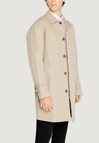 Cappotto impermeabile beige con chiusura a bottoni, ampio colletto e tasche laterali; presenta maniche larghe e una texture liscia e leggera.