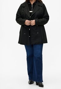Svart dubbelknäppt trenchcoat med stora knappar, lager-på-lager över en randig topp. Matchas med utsvängda blå jeans och svarta klackar.