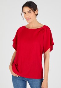Blusa rossa a maniche corte con tessuto testurizzato e dettaglio arricciato sulla spalla sinistra. Vestibilità comoda, scollatura rotonda, abbinata a denim.