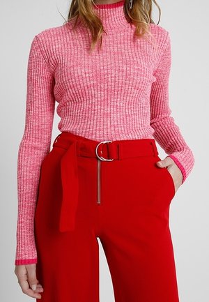 Gestreifter, hochgeschlossener rosa Pullover mit langen Ärmeln kombiniert mit leuchtend roten, weit geschnittenen Hosen, die einen Gürtel und Reißverschlussdetails aufweisen.