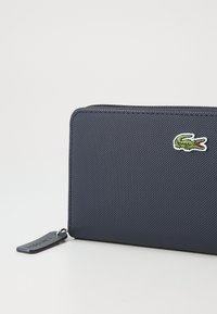 Marine blødt pung med tekstureret stof, der har et grønt krokodillelogo broderet og et sort mærke med brandnavnet. Glat, kompakt design.