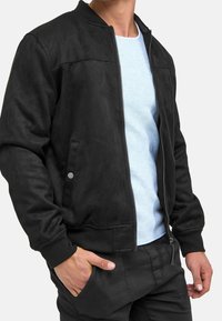 Zwarte bomberjack van zacht suede met een rits aan de voorkant, geribde manchetten en twee zijzakken. Gecombineerd met een lichtblauw shirt eronder.
