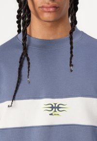 Sudadera de manga larga azul y blanca con un logo geométrico amarillo en el pecho, hecha de una tela texturizada y con un corte relajado.