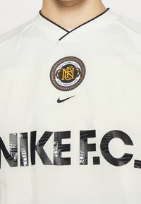 Biała koszulka piłkarska Nike z czarną okrągłą naszywką klubową, wyraźnym czarnym napisem "NIKE F.C." oraz dekoltem w serek z kontrastowym kołnierzykiem.