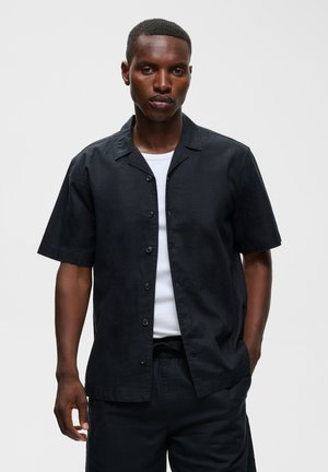 Jeune homme portant une chemise noire à manches courtes boutonnée par-dessus un t-shirt blanc, debout avec une main dans la poche sur un fond uni.