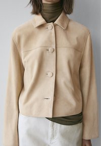 Beige ruskindjakke med krave, enkelt række knapper og en cropped pasform. Kombineret med en mørkegrøn rullekrave under.