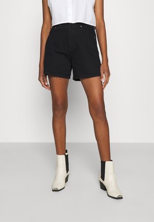 Donna che indossa pantaloncini neri a vita alta, camicetta bianca corta senza maniche e stivaletti bianchi con lati elastici neri, in piedi su pavimento grigio.