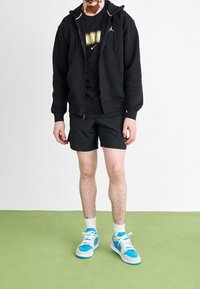 Person wearing black zip-up hoodie, black shorts, white socks, and blue and white sneakers standing on green floor against white wall.