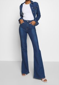 Jeansjacka i mörkblått med fickor och knäppning, kombinerad med högmidjade, utsvängda mörkblå jeans, bärs med vita klacksandaler.