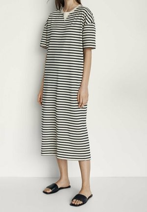 Femme portant une robe midi à rayures horizontales noires et blanches avec des manches courtes et des sandales plates noires, debout devant un fond uni.
