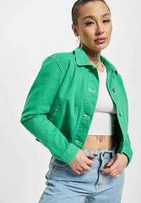 Joven con tatuaje en el cuello que lleva una chaqueta verde, top corto blanco y vaqueros azul claro, posando con las manos cerca de la cintura.