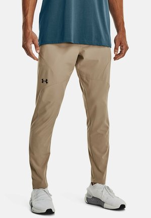 Mann trägt hellbraune Under Armour Hose, weiße Sportschuhe und blaues Hemd, steht mit entspannten Händen an den Seiten.