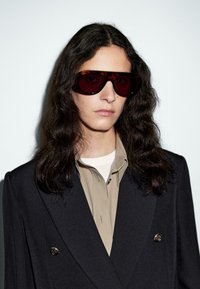 Person mit langen, dunklen, gewellten Haaren, die übergroße Schildpatt-Sonnenbrille, ein beiges Hemd mit Kragen und ein schwarzes, doppelt knöpfbares Sakko trägt, vor einer weißen Wand.