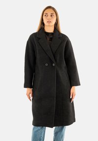 Donna che indossa un lungo cappotto nero doppiopetto sopra jeans blu, in piedi davanti a uno sfondo chiaro e guardando di lato.