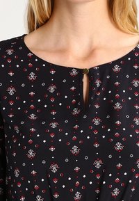 Svart blus med ett blommigt mönster i rött och vitt. Rund halsringning med en nyckelhål-detail fram och en liten metallisk knappaccent.