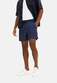 Costumini da bagno blu navy con vita elastica, lacci e tasche laterali, abbinati a una camicia a maniche corte con bottoni e sneakers bianche.
