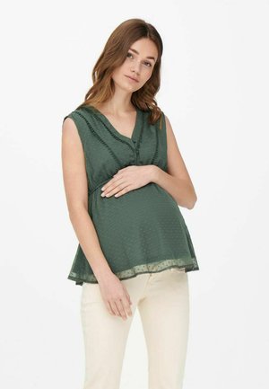 Maglietta premaman verde con scollatura a V, maniche corte, tessuto a pois texturizzato e orlo fluido, indossata sopra pantaloni color crema.