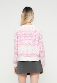 Woman with long blonde hair wears white and pink patterned sweater and gray mini skirt, standing facing away against plain background.