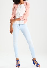 Veste bomber rose clair sur un top peplum blanc avec des détails en œillets, associée à un jean skinny bleu clair et des talons bleus à brides.