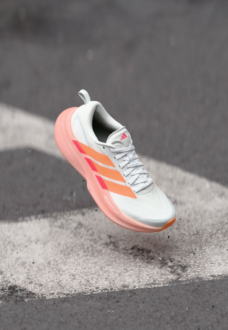 Chaussure de course avec une tige blanche, des accents orange et rose, et une semelle rose amortie. Dispose de trois bandes parallèles sur le côté.