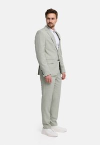 Man standing facing sideways in a light gray suit, white dress shirt, and white sneakers against a plain white background.