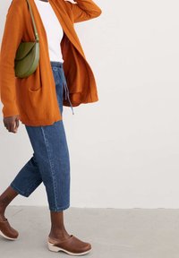 Bränd orange cardigan med öppen framsida, kombinerad med ljusa denimankelbyxor och bruna träklackar. Har en grön axelväska.