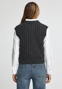 Grå stickad väst med snöre-mönster, ärmlös design, vågig nederkant, i kombination med en vit långärmad skjorta och denimjeans. Bakifrån.