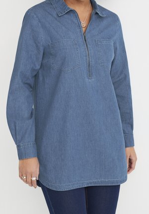 Kobieta nosząca długą niebieską tunikę jeansową z zamkiem z przodu i dwoma kieszonkami na piersi, w połączeniu z ciemnoniebieskimi spodniami.