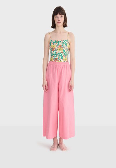 Jeune femme debout, portant un caraco à motif floral, un pantalon ample taille haute rose, et des chaussures transparentes rose à enfiler.