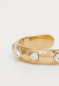 Aranyszínű karperec emelt kör alakú díszekkel, melyekben átlátszó műgemstone-ok vannak, sima felülettel és lekerekített élekkel.