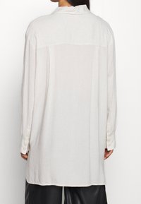 Camicia di lino crema con maniche lunghe, un giromanica sul retro e polsini abbottonati; presenta una vestibilità rilassata e una sottile trama.
