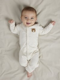 Krämfärgad babybody med broderat björnansikte på br östet, tillverkad av mjukt tyg. Bebisen ligger på en ljusbeige yta.
