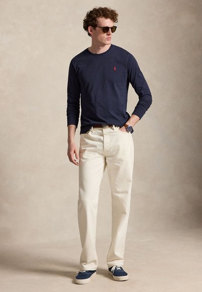 Férfi tengerészkék hosszú ujjú ingben, krémszínű nadrágban, kék tornacipőben, napszemüvegben és karórával, semleges háttér előtt áll.