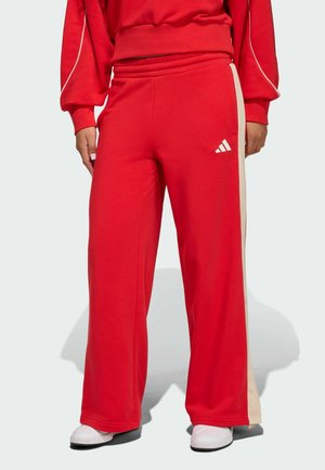 Rote weit geschnittene Hose mit beigen Seitenstreifen und einem weißen Adidas-Logo, getragen mit weißen Sneakers und einem passenden roten Sweatshirt.