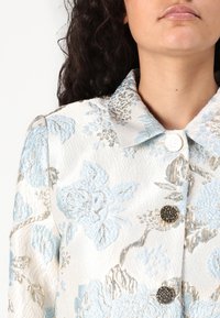 Valkoinen kukkajacquard-takki, jossa on vaaleansinisiä ja metallinhohtoisia hopeisia yksityiskohtia. Takissa on napitettu etuosa ja jämäkkä kaulus.