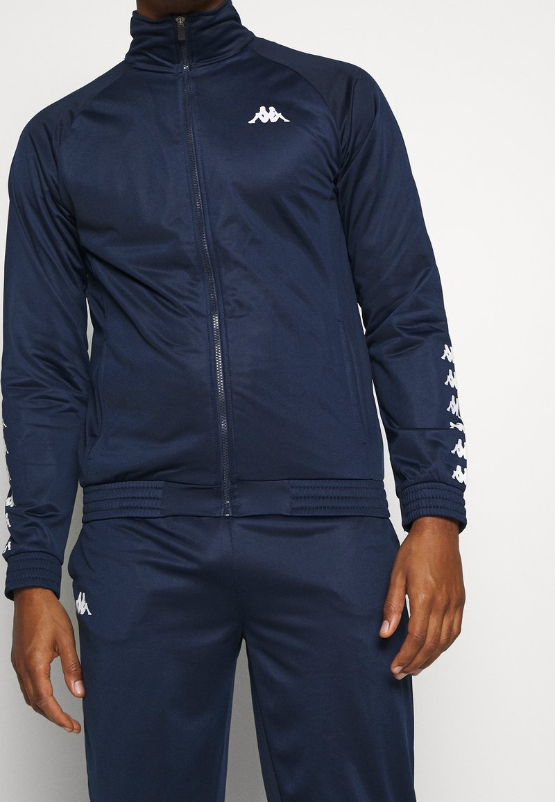 Homme portant une veste de sport bleu marine zippée et un pantalon assorti avec des logos blancs sur la poitrine, les manches et la cuisse.