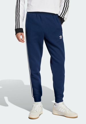 Mann trägt dunkelblaue Adidas-Trainingshose mit weißen Seitenstreifen und weiße Sneakers, steht vor hellem grauem Hintergrund.