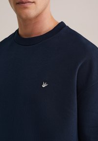 Temno modra jopa z rebrastim izrezom, z majhnim vezenim logotipom ptice na prsih. Mehka, gladka tekstura.