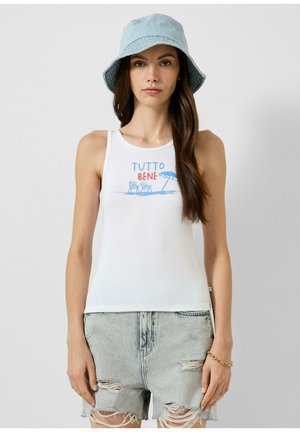 Femme portant un chapeau bob bleu clair, un débardeur blanc avec l'inscription "Tutto Bene" et un motif plage, et un short en jeans clair déchiré avec un bracelet en or.