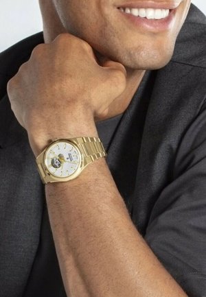 Montre en acier inoxydable doré avec un cadran blanc, des accents noirs et dorés, et un bracelet métallique. Engrenages visibles à travers un dos de boîtier transparent.