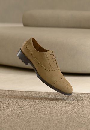 Svetlohnedá semišová oxfordská topánka s brogue detailmi, vznášajúca sa nad texturovaným béžovým kobercom pred béžovou pohovkou.