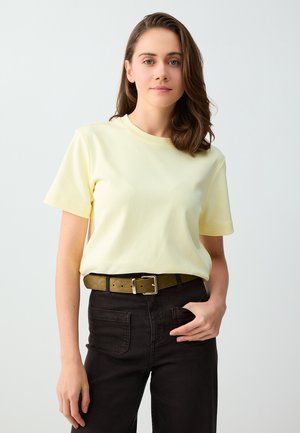 Giovane donna con capelli castani fino alle spalle che indossa una maglietta giallo pallido infilata in pantaloni neri con una cintura verde, in piedi davanti a uno sfondo semplice.