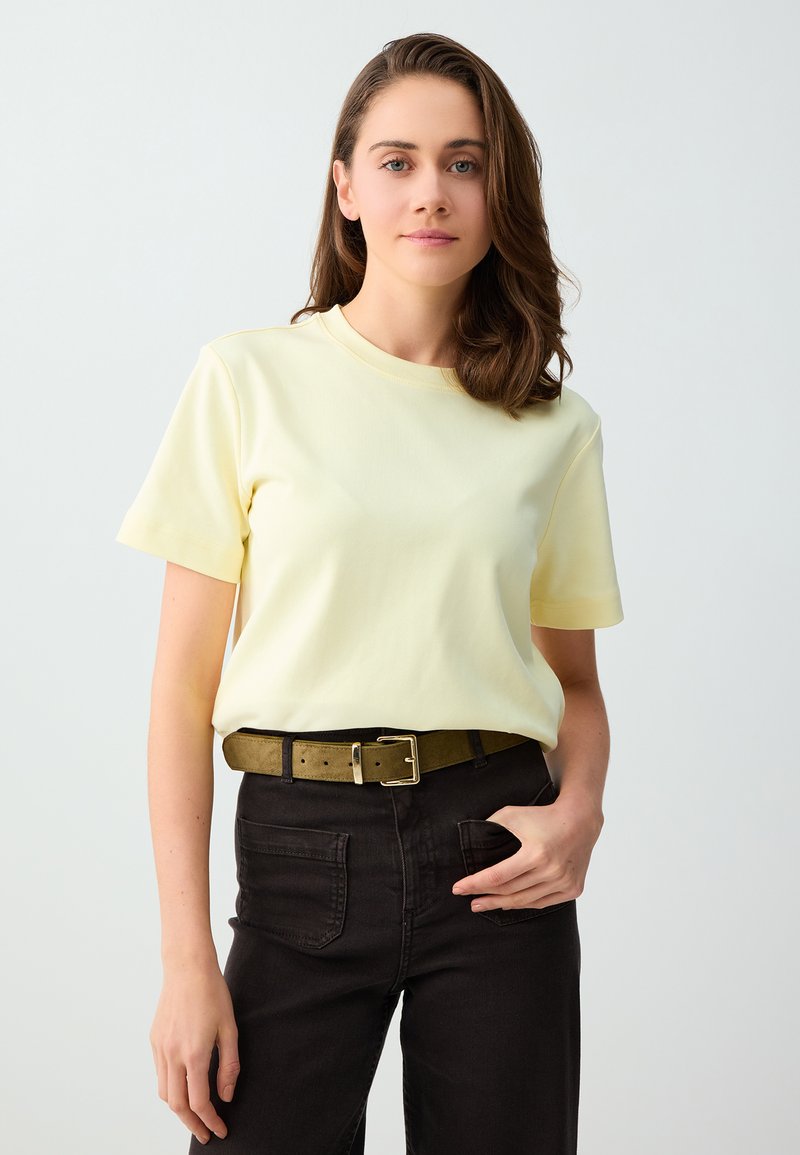 Jeune femme aux cheveux bruns mi-longs, portant un T-shirt jaune pâle rentré dans un pantalon noir avec une ceinture verte, debout devant un fond uni.