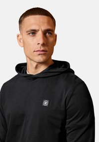 Black hoodie made of cotton blend material, featuring a drawstring hood, a front logo patch, and a fitted design. Smooth texture.
