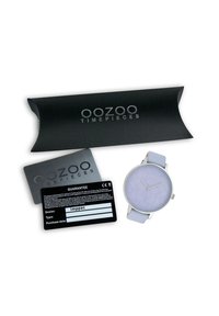 Montre lilas avec un cadran rond texturé, lunette argentée et bracelet lavande. Livrée avec un emballage noir et une carte de garantie.